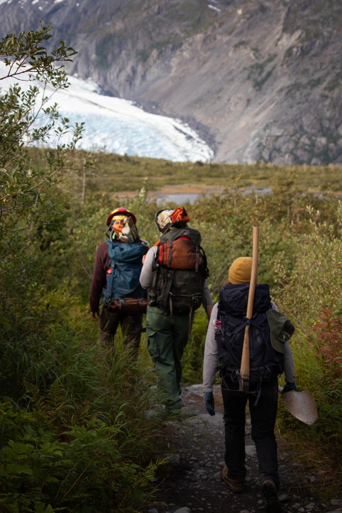 Three trail workers with tools strapped to their backpacks walk down a trail towards a large glacier. 